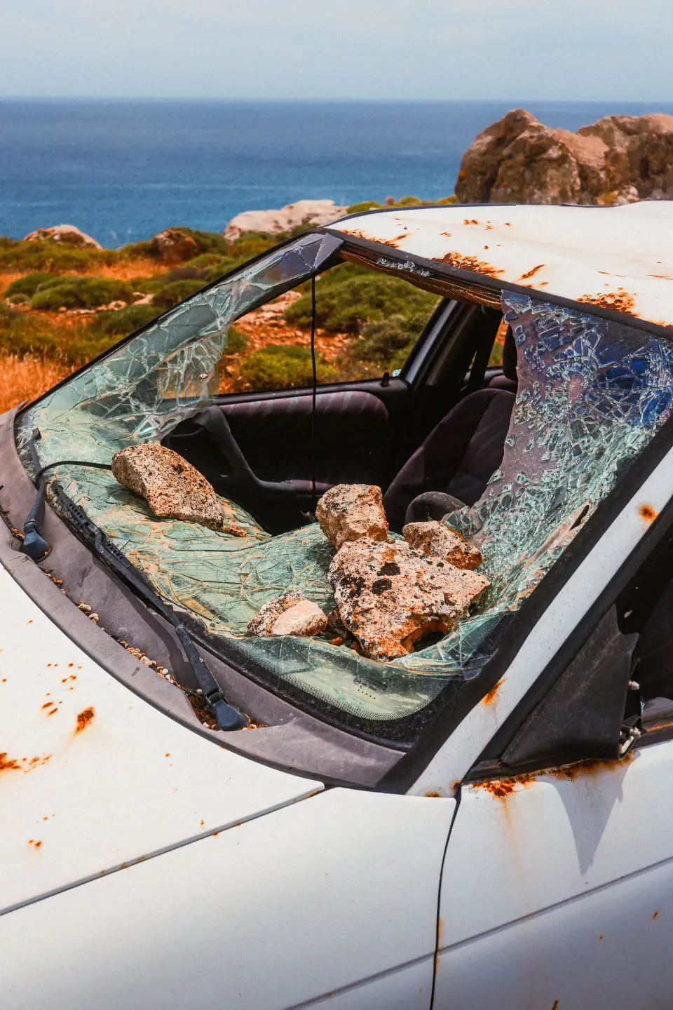 A car by the sea with its windshield caved in by rocks, glass spidered across the dash